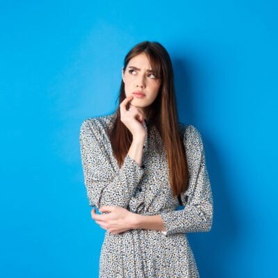 Thoughtful serious woman in dress looking away pensive, standing hesitant and unsure, thinking on blue background.